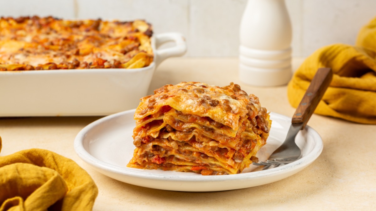 A slice of lentil lasagna on a plate. In the background, the rest can be seen in a baking dish.