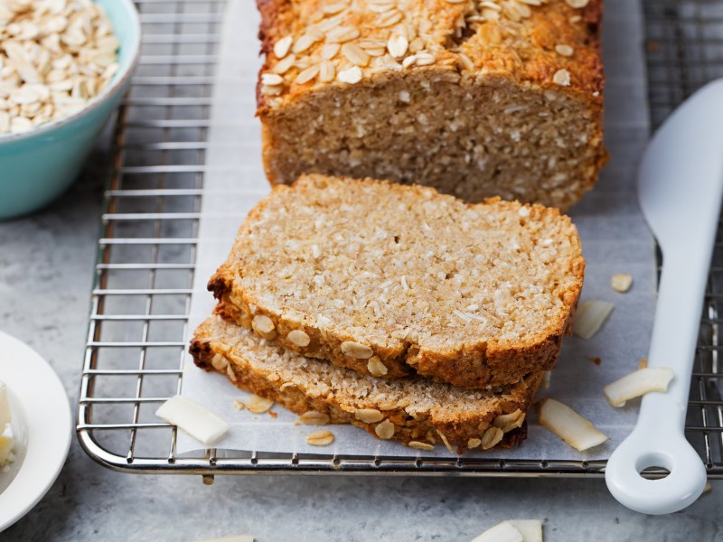 Sliced oatmeal quark bread on a board, next to it a bowl of oatmeal and a knife.