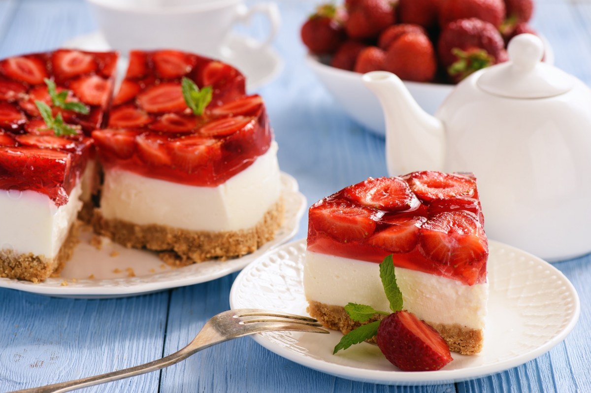 A piece of no-bake strawberry cheesecake on a plate. In the background, the rest of the cake, a teapot and a bowl of strawberries can be seen blurred.