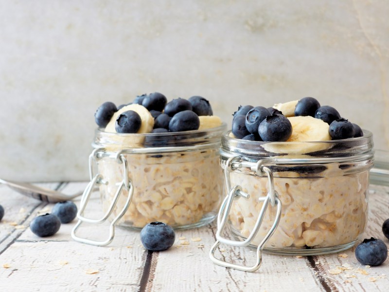 2 glasses of overnight oats garnished with blueberries and banana slices, with some blueberries scattered around.