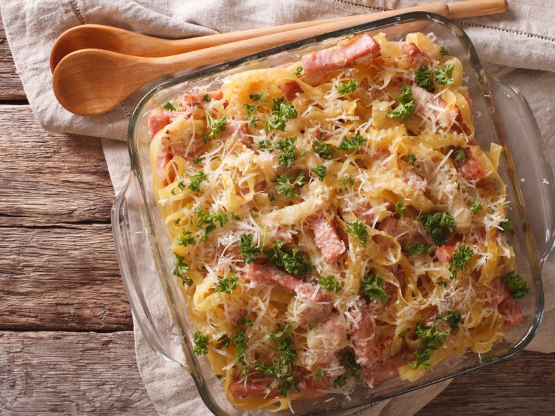 A noodle and ham gratin in a glass baking dish stands on a cloth towel. Wooden cutlery lies on top.