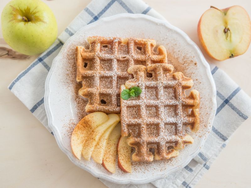 Oatmeal-applesauce waffles arranged on a plate with apple slices and powdered sugar. The plate is on a kitchen towel. Two apples lie next to the plate as decoration.