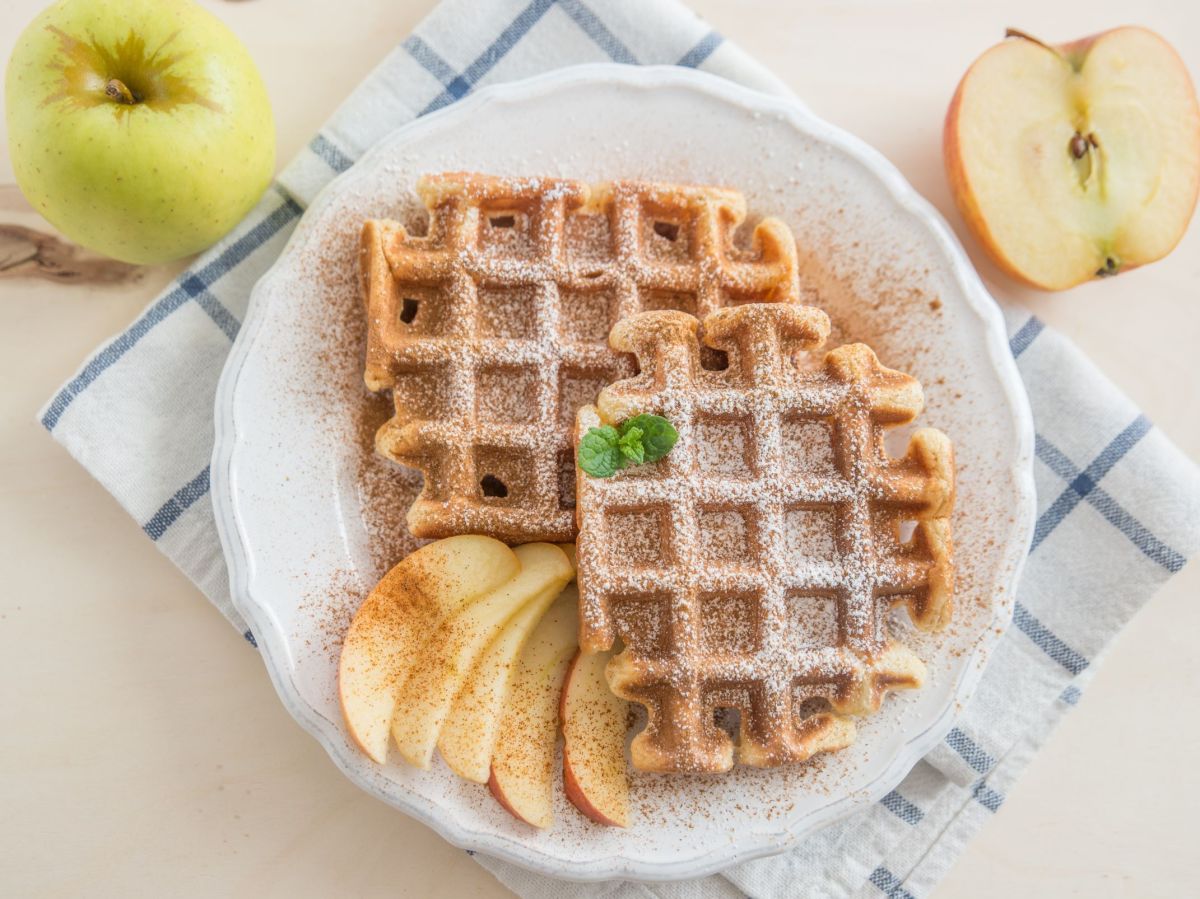Oatmeal-applesauce waffles arranged on a plate with apple slices and powdered sugar. The plate is on a kitchen towel. Two apples lie next to the plate as decoration.