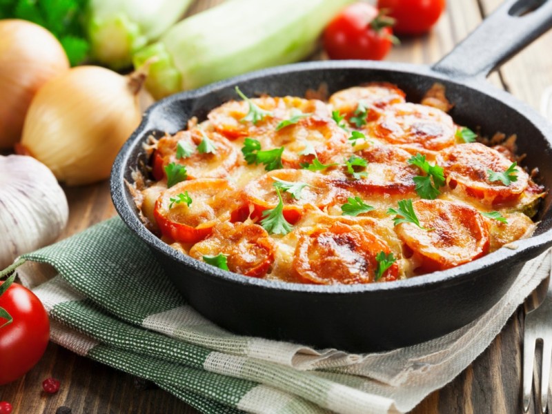 Tomato and zucchini casserole in an ovenproof pan on a dish towel. Fresh ingredients lie in the background.