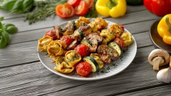 A plate of grilled tortelloni skewers. The plate is on a wooden surface. Yellow bell peppers, tomatoes, and fresh herbs are scattered around.