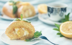 A slice of lemon swiss roll, decorated with mint, on a plate. A cup of coffee and the rest of the cake can be seen in the background.